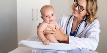 A baby boy sitting on scale during weight measuring exam at doctor office. Pediatrician weighting baby girl on scales