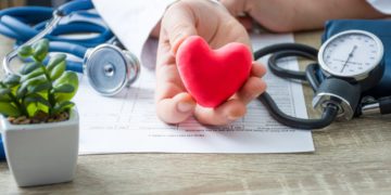 Doctor of internal medicine and cardiologist holding in his hands and shows to patient figure of red card heart during medical consultation. Explanation of causes of heart, diagnosis and treatment