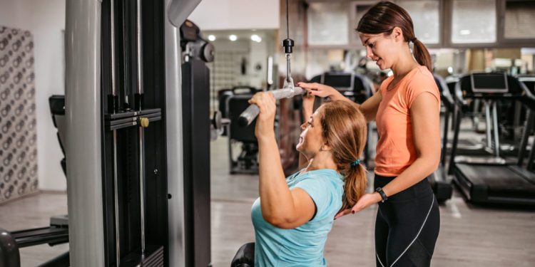 Senior woman doing lat pulldowns exercise in gym with trainers help