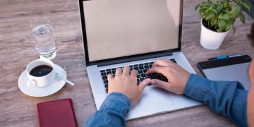 A man sits at his desk with his laptop, coffee, water, and flowers.