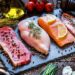 High angle view of four different types of animal protein like a raw beef steak, a raw chicken breast, a raw salmon fillet and a raw pork steak on a stone tray surrounded by condiments, spices and vegetables
