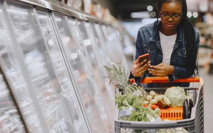 Woman in a grocery store using smartphone while pushing a shopping cart filled with groceries.