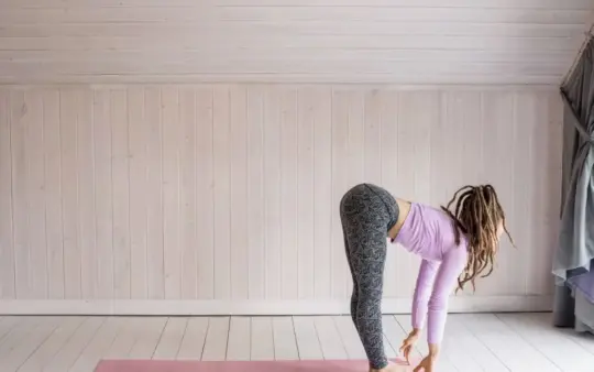 Woman with dreadlocks performs a forward bend yoga pose on a pink mat indoors.