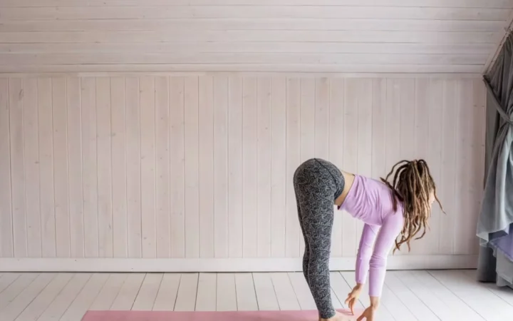 Woman with dreadlocks performs a forward bend yoga pose on a pink mat indoors.