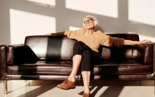 Elderly woman with gray hair and eyeglasses relaxing on a leather sofa in a bright room.