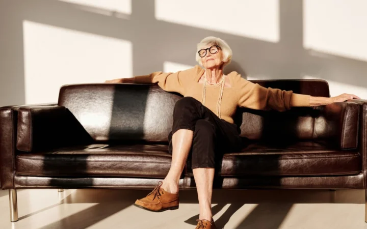 Elderly woman with gray hair and eyeglasses relaxing on a leather sofa in a bright room.