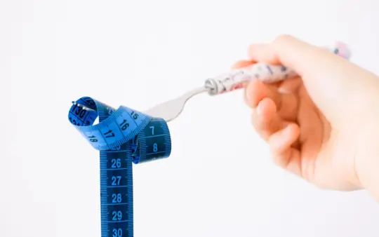 Hand holding a fork wrapped with a blue measuring tape suggesting diet or weight management.