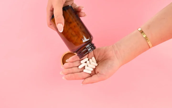 Close-up of two hands pouring white capsules from a bottle on a bright pink background.
