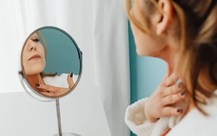 Woman in a bathrobe applying skincare while looking in the mirror.