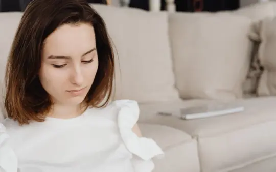 Young woman in a white top sits on a beige couch looking down, creating a serene indoor scene.