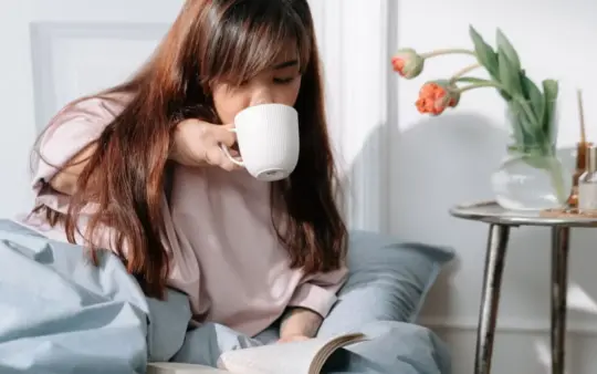 Asian woman enjoying her morning with coffee and a book in a cozy bedroom setting.