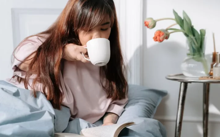 Asian woman enjoying her morning with coffee and a book in a cozy bedroom setting.