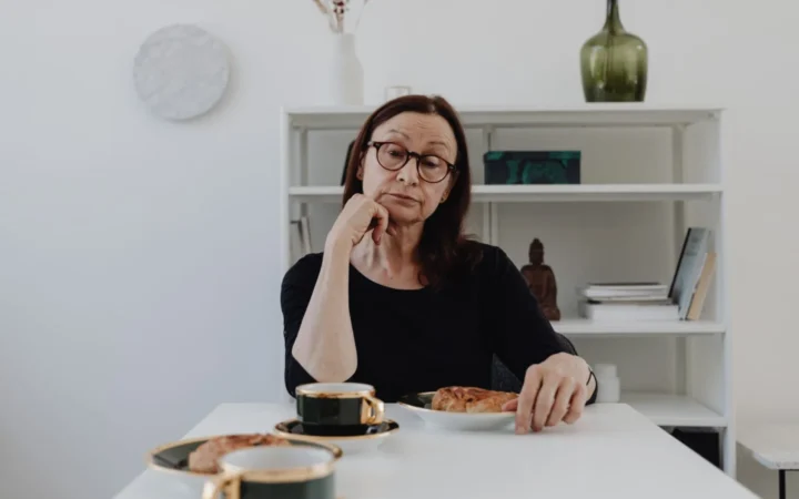 Elderly woman sits pensively at a white table with coffee and pastries.