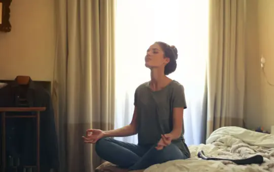 Adult woman practicing meditation on her bed surrounded by a calm bedroom atmosphere.