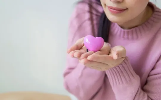 Woman in a pink sweater holding a pink heart-shaped toy, symbolizing love and kindness.
