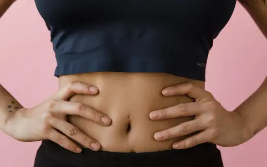 Close-up shot of a woman's toned stomach with hands on hips, against a pink background.