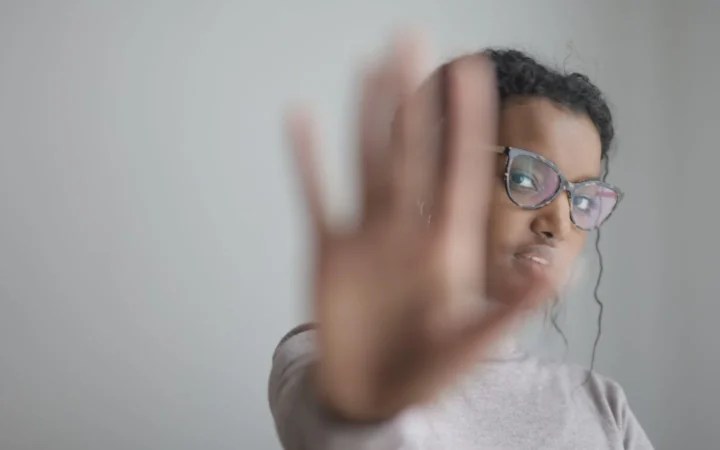 Young African American female wearing casual clothes and eyeglasses outstretching arm at camera while standing near grey wall