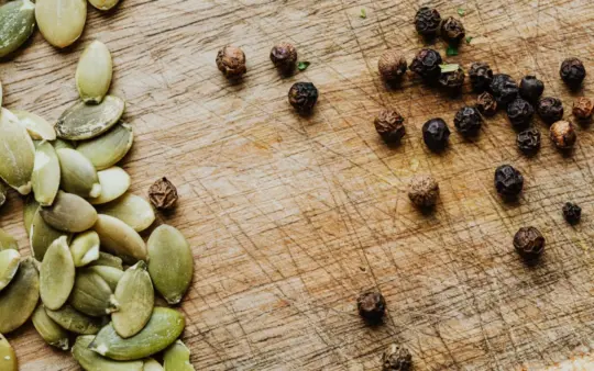 Close-up of pumpkin seeds and black peppercorns on a wooden cutting board.