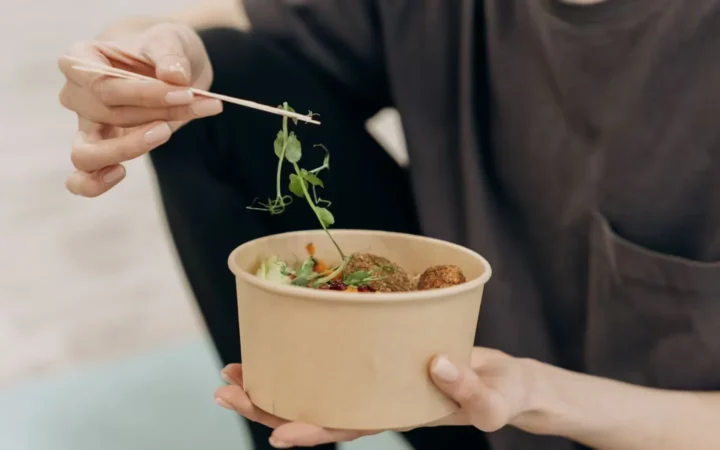 Woman holding a paper bowl filled with a healthy vegetarian meal in an indoor setting, promoting wellness.