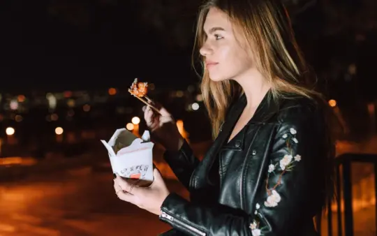 Young woman eating Asian takeout at night, city lights glowing in the background.
