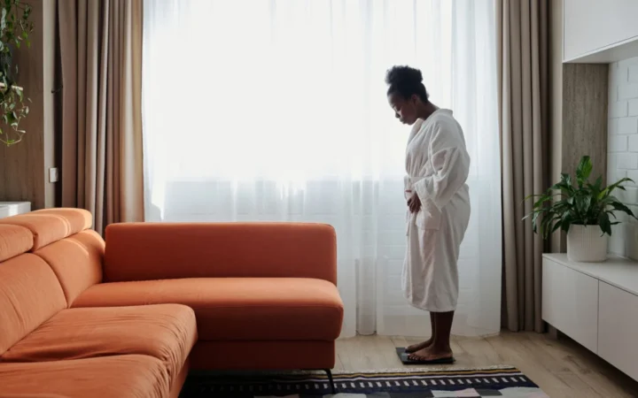 African American woman in a bathrobe checks her weight on a scale near an orange sofa.