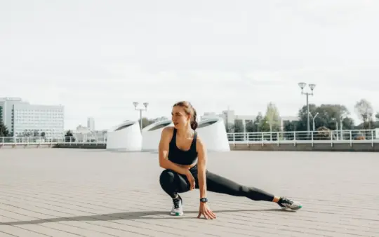 Young woman in black sportswear stretching outdoors in a sunny urban environment.