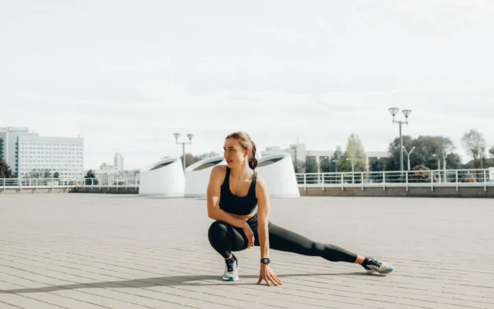 Young woman in black sportswear stretching outdoors in a sunny urban environment.