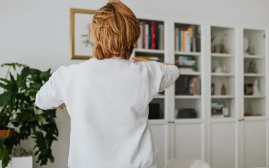 A woman in a white sweatshirt reaching towards her lower back in a bright living room