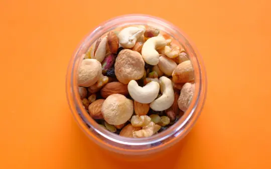 Close-up of a mix of nuts in a clear container against an orange background.