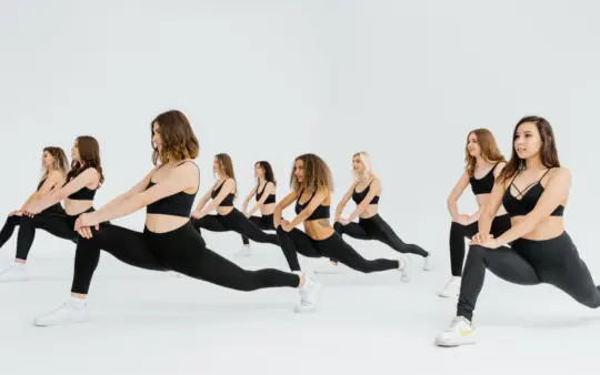 Group of women in black activewear performing lunges in a bright indoor studio, promoting fitness.