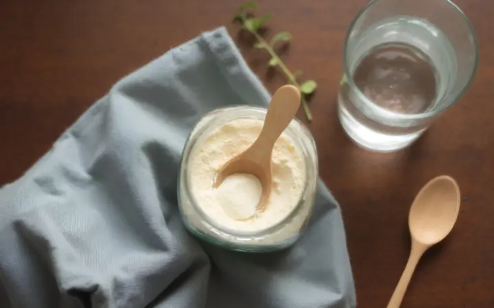 Collagen peptides for skin: a glass jar of unflavoured collagen powder with a small wooden scoop resting in it, a tall glass of clear water filled two thirds, and a teaspoon, on a dark walnut kitchen table with the rest of the table bare, seen from directly above, generated illustration