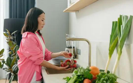 Asian woman washes strawberries at kitchen sink, promoting healthy living.