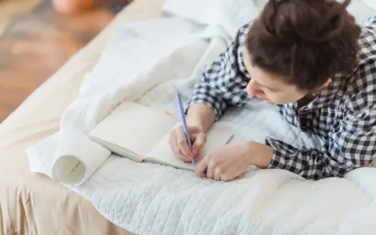 Woman writes in a journal while relaxing on a cozy bed with soft lighting and peaceful surroundings.