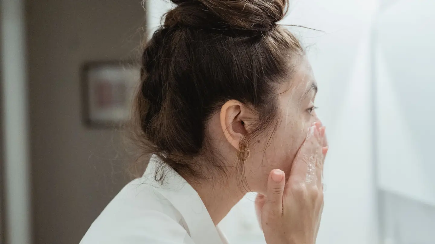 Best face wash for sensitive oily skin: a young woman in a bathrobe follows her skincare routine, washing her face in a