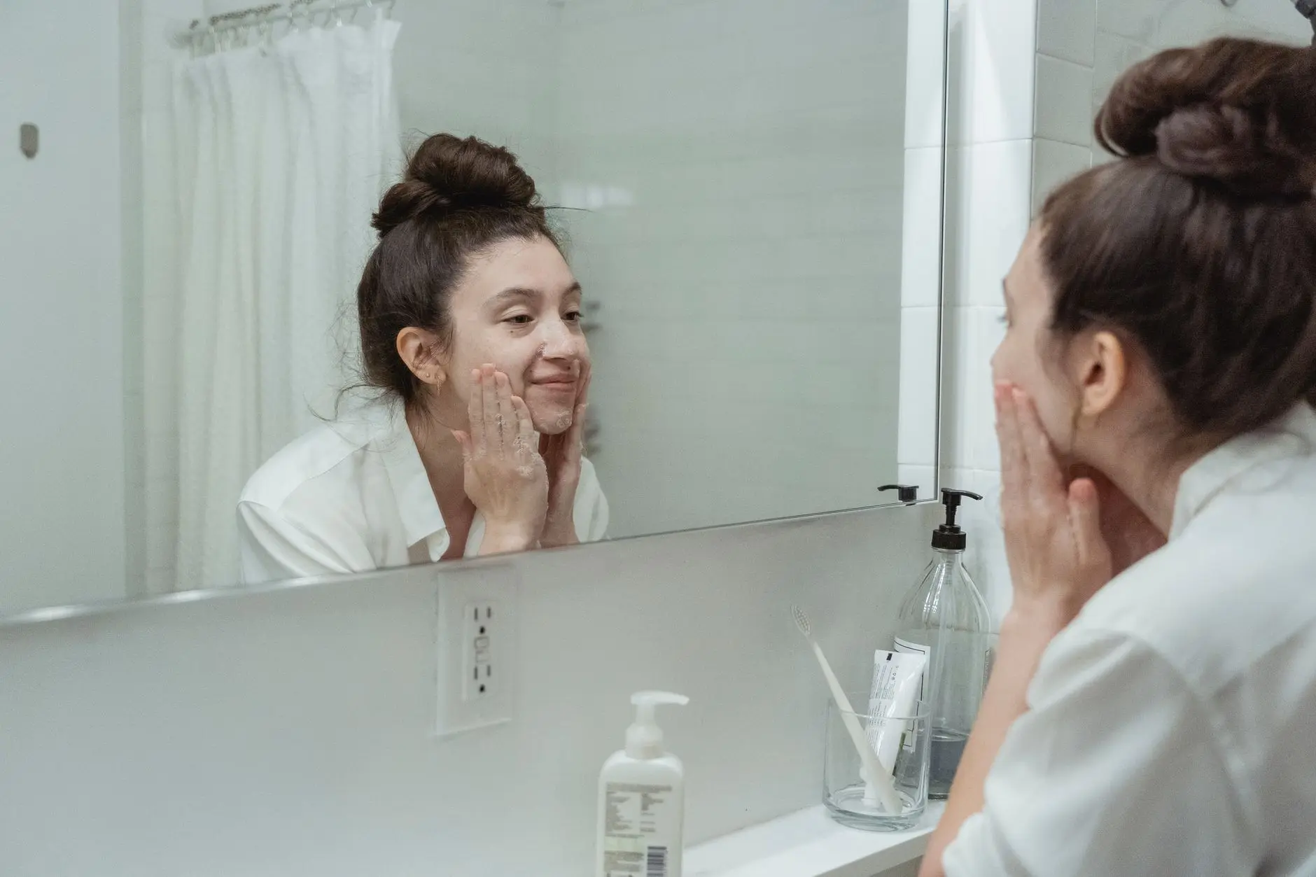 Woman performing skincare routine in a bathroom, applying face cream while looking into a mirror