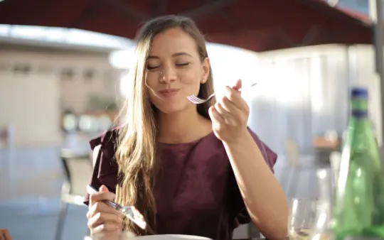Woman savoring her meal with a smile, dining outdoors in a pleasant setting.
