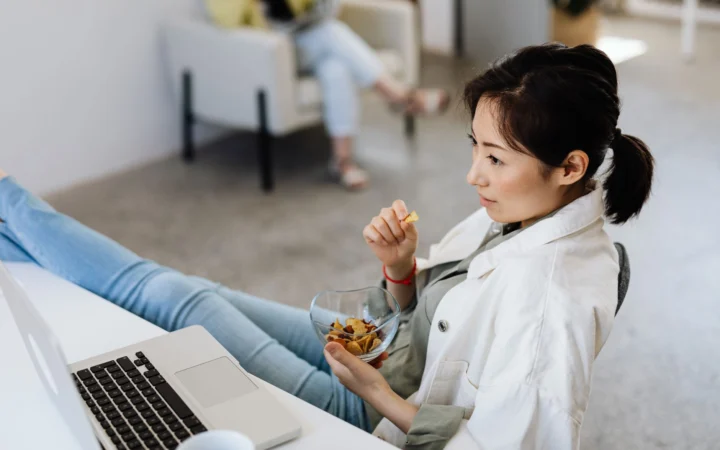 Young woman enjoying a snack while working on a laptop in a casual office setting.