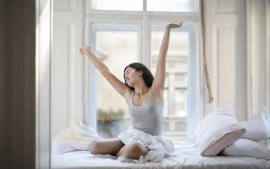 Woman in a gray tank top stretches joyfully on her bed in a bright room, embracing a new day.