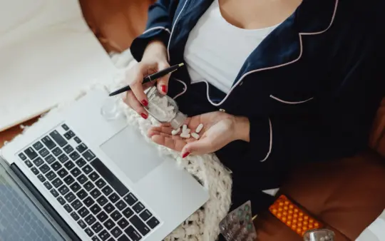 Close-up of a woman in pajamas holding pills next to a laptop, illustrating remote work and health care at home.