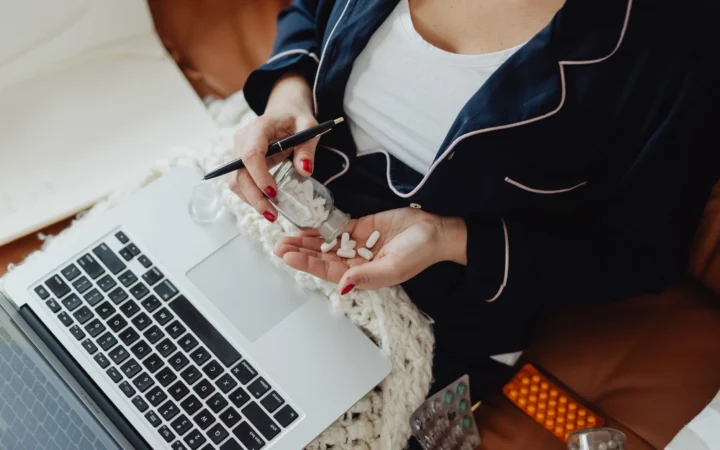 Close-up of a woman in pajamas holding pills next to a laptop, illustrating remote work and health care at home.