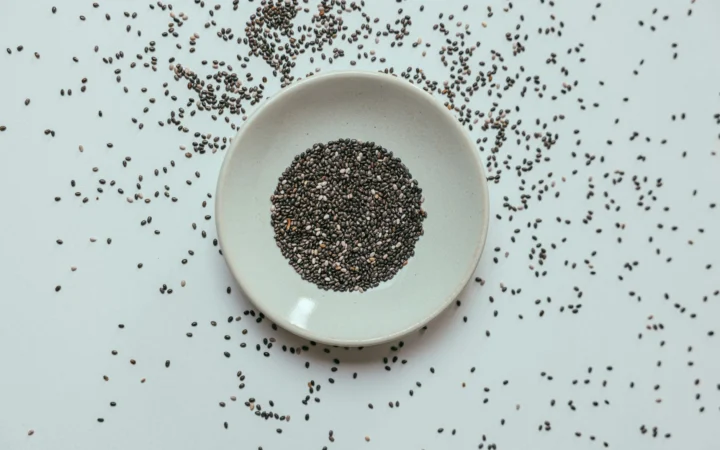 Top view of chia seeds in a white bowl with scattered seeds around on a flat surface.