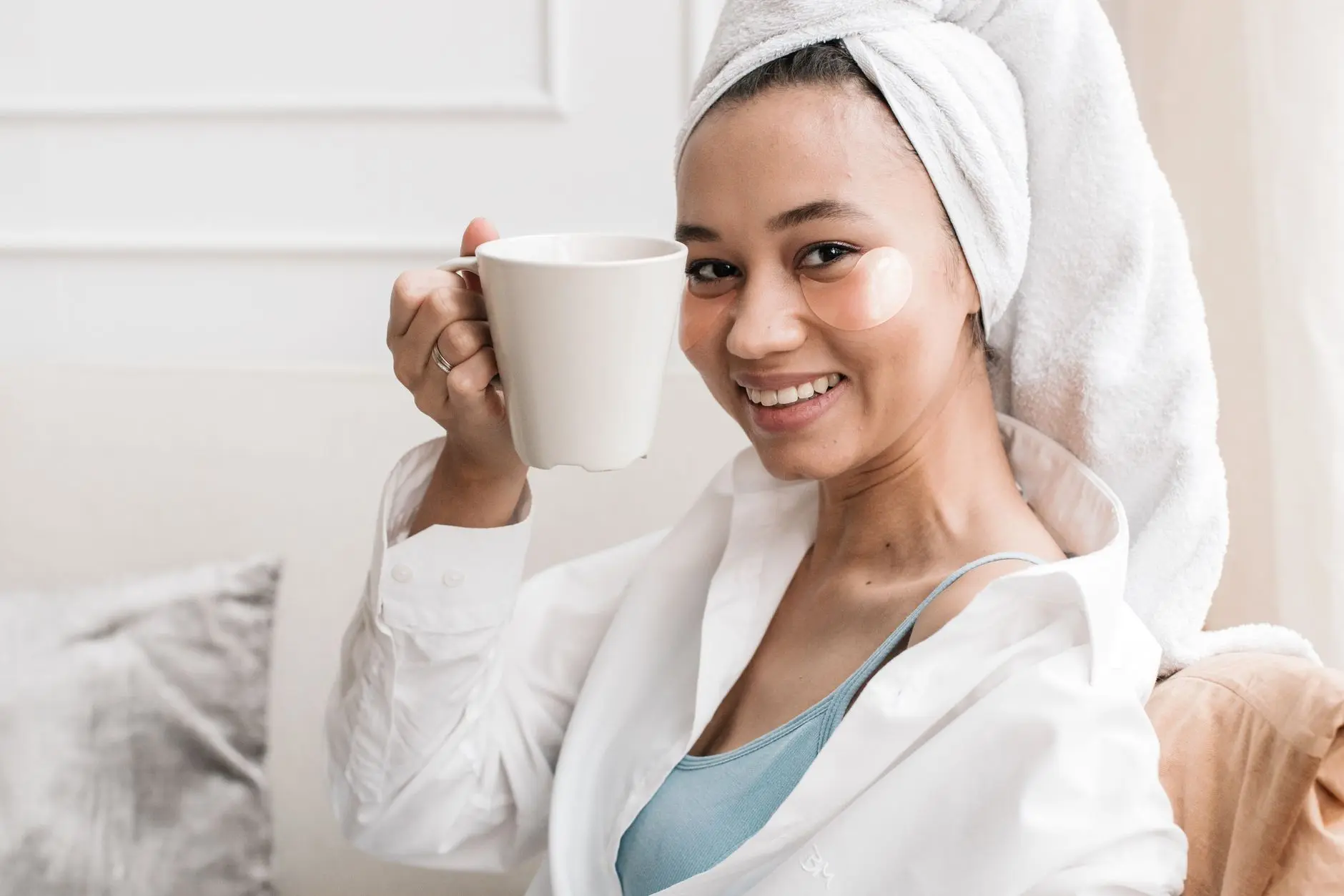 An Asian woman with eye patches and a towel turban enjoys a hot drink indoors, emphasizing beauty and relaxation