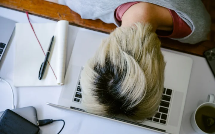 From above of tired fatigued unrecognizable female in casual wear resting head on netbook keyboard near notebook and cup of