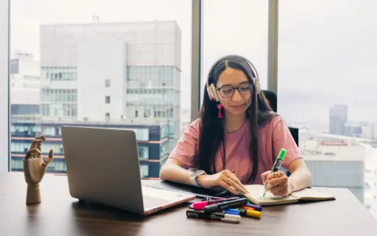 Young woman wearing headphones, writing in notebook at office desk with city view.