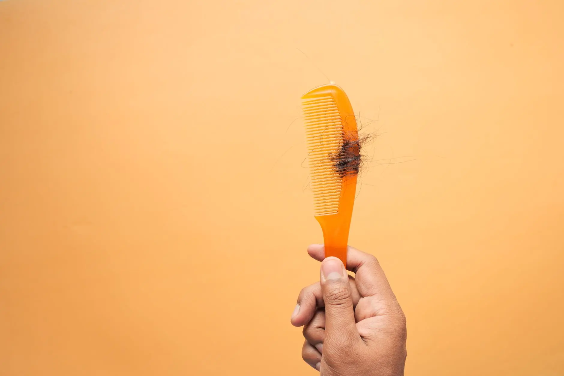 Close-up of hand holding a comb with hair against an orange backdrop, highlighting hair loss concept