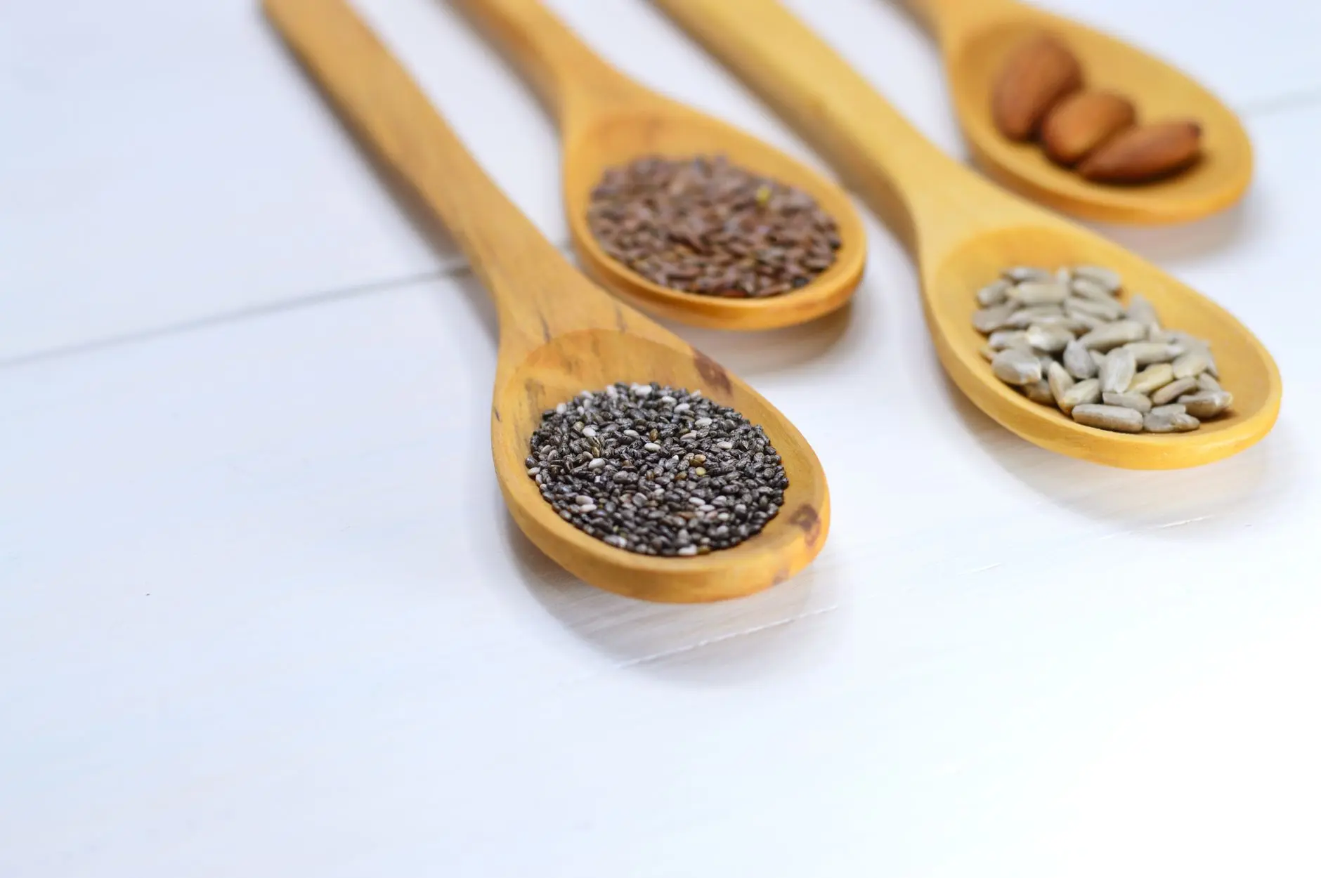 A variety of seeds and nuts in wooden spoons displayed on a white surface