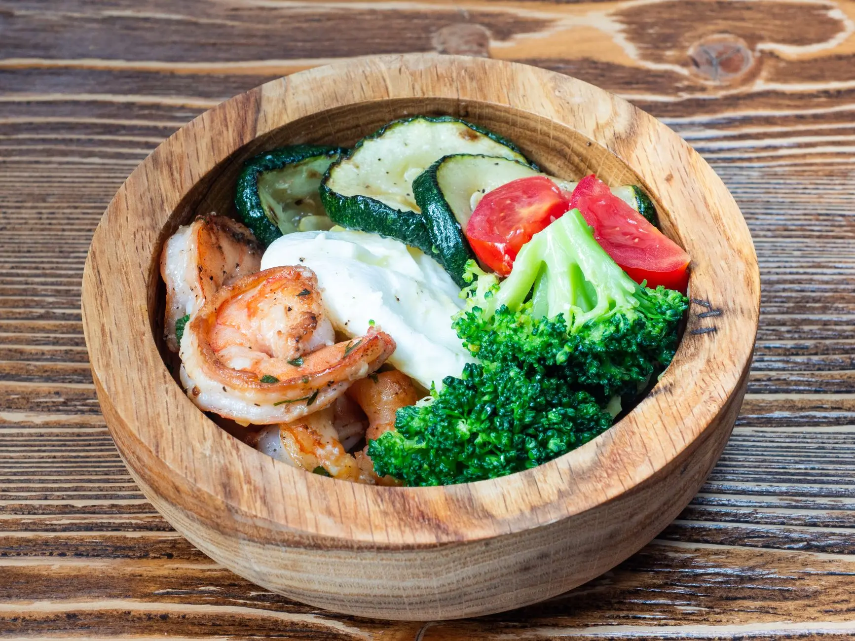 A wooden bowl filled with shrimps, broccoli, zucchini, and tomatoes on a rustic table