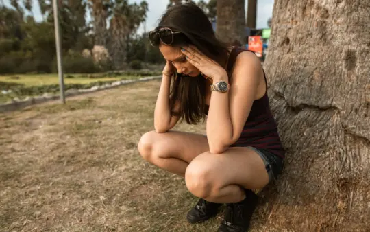 Woman kneels by a tree holding her head, expressing stress or anxiety in an outdoor park.