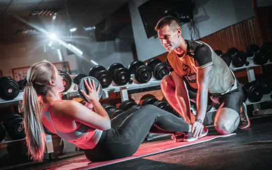 Personal trainer assists a woman doing sit-ups with a medicine ball in a gym setting.