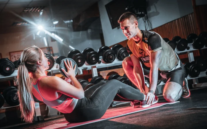Personal trainer assists a woman doing sit-ups with a medicine ball in a gym setting.